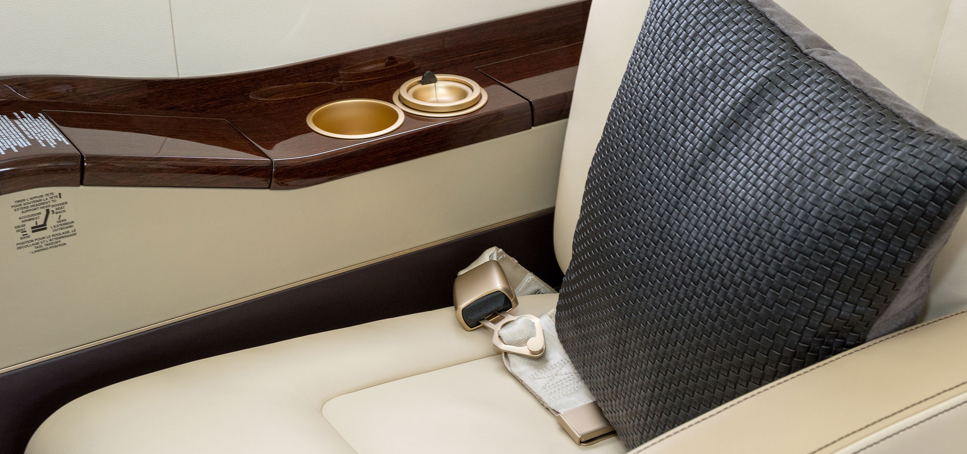 A woven pillow rests against a first-class airplane seat made of leather and polished wood with brushed gold fixtures